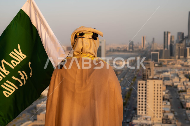 توحيد الوطن وتأسيس السعودية في 1727م, ذكرى يوم التأسيس في 22 فبراير, مؤسس الدولة السعودية الاولى الامام محمد بن سعود, صورة مقربة من الخلف لرجل عربي خليجي سعودي يرتدي البشت والغترة يحمل بيده علم التأسيس, الزي التقليدي ليوم التأسيس, خلفية احياء مدينة الرياض.