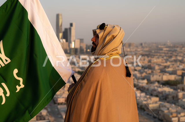 ذكرى يوم التأسيس في 22 فبراير، توحيد البلاد وتأسيس السعودية في 1727م, مؤسس الدولة السعودية الاولى الامام محمد بن سعود, صورة مقربة من الخلف لرجل عربي خليجي سعودي يرتدي البشت والغترة يحمل بيده علم التأسيس, الزي التقليدي ليوم التأسيس, خلفية احياء مدينة الرياض.