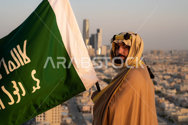 الاحتفال بذكرى يوم التأسيس في 22 فبراير، مؤسس الدولة السعودية الاولى الامام محمد بن سعود, تاريخ توحيد البلاد وتأسيس السعودية في 1727م, صورة مقربة لرجل عربي خليجي سعودي يرتدي البشت والغترة يحمل بيده علم التأسيس, الزي الشعبي للمناسبات الوطنية, خلفية مدينة الرياض.