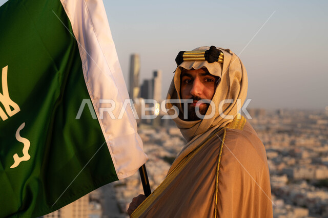 مؤسس الدولة السعودية الاولى الامام محمد بن سعود, تاريخ توحيد البلاد وتأسيس السعودية في 1727م, صورة مقربة لرجل عربي خليجي سعودي يرتدي البشت والغترة يحمل بيده علم التأسيس, الزي الشعبي للمناسبات الوطنية, الاحتفال بذكرى يوم التأسيس في 22 فبراير، خلفية مدينة الرياض.