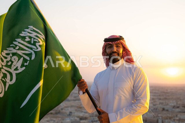 تعزيز الهوية الوطنية واظهار الاخلاص للبلاد، رجل عربي خليجي سعودي مبتسم يرتدي الشماغ والزي التقليدي يحمل بيده علم المملكه  في وضح النهار، يوم العلم 11 مارس، الاحتفال بذكرى اليوم الوطني السعودي 23 مارس، أعياد ومناسبات شعبية، خلفية أحياء ومباني مدينة الرياض.