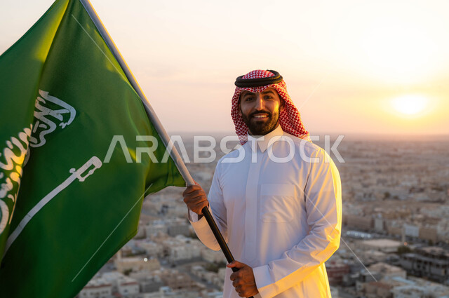 تعزيز الروح الوطنية واظهار الاخلاص للبلاد، رجل عربي خليجي سعودي مبتسم يرتدي الشماغ والزي التقليدي يحمل بيده علم المملكه  في وضح النهار، يوم العلم 11 مارس، الاحتفال بذكرى اليوم الوطني السعودي 23 مارس، أعياد ومناسبات شعبية، خلفية أحياء ومباني مدينة الرياض.