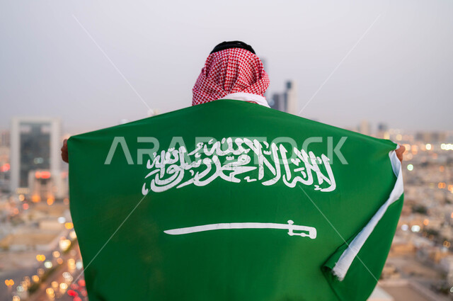 احتفالات ومناسبات شعبية وطنية، اظهار الحب والولاء للملكه، احياء ذكرى اليوم الوطني السعودي 23 سبتمبر، صورة مقربة من الخلف لرجل عربي خليجي سعودي يرتدي الزي التقليدي ويضع علم السعودية على كتفيه في العيد الوطني، يوم العلم 11 مارس ،خلفية أبراج ومباني مدينة الرياض.