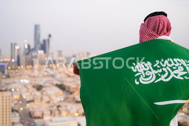 احتفالات ومناسبات وطنية شعبية، اظهار الحب والولاء للملكه، احياء ذكرى اليوم الوطني السعودي 23 سبتمبر، صورة مقربة من الخلف لرجل عربي خليجي سعودي يرتدي الزي التقليدي ويضع علم السعودية على كتفيه في العيد الوطني، يوم العلم 11 مارس ،خلفية أبراج ومباني مدينة الرياض.