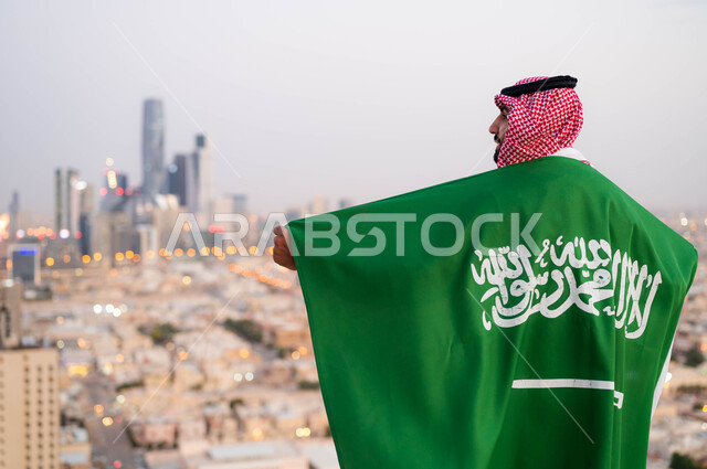 مناسبات واحتفالات وطنية شعبية، اظهار الحب والولاء للملكه، احياء ذكرى اليوم الوطني السعودي 23 سبتمبر، صورة من الخلف لرجل عربي خليجي سعودي يرتدي الزي التقليدي ويضع علم السعودية على كتفيه في العيد الوطني، يوم العلم 11 مارس ،خلفية أبراج ومباني مدينة الرياض.