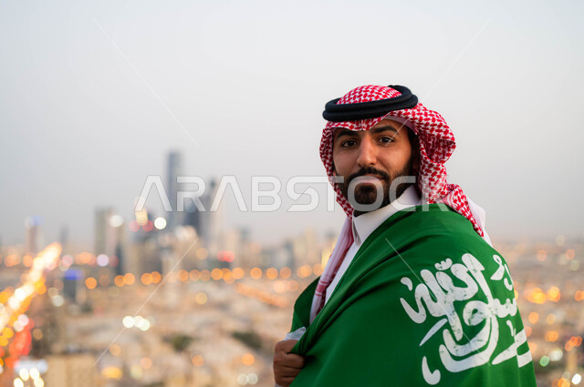  احياء ذكرى اليوم الوطني السعودي 23 سبتمبر، مناسبات واحتفالات وطنية شعبية، رجل عربي خليجي سعودي مبتسم يرتدي الثوب و الشماغ في ويضع علم السعودية على كتفيه في العيد الوطني، يوم العلم 11 مارس، النظر إلى الكاميرا، خلفية أبراج ومباني مدينة الرياض.