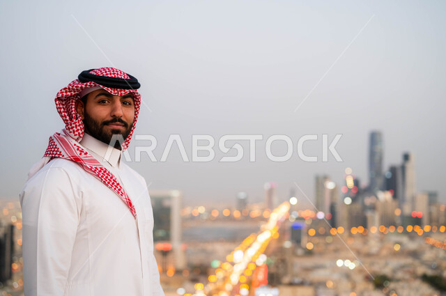  التقدم والتطور العمراني في المدينة، الاعتزاز والفخر بالنمو المعماري في السعودية، التجول والتنزه واستنشاق الهواء النقي في الخارج، صورة مقربة لرجل عربي خليجي سعودي مبتسم يرتدي الشماغ و الزي السعودي التقليدي ينظر الى الكاميرا، خلفية احياء مدينة الرياض.