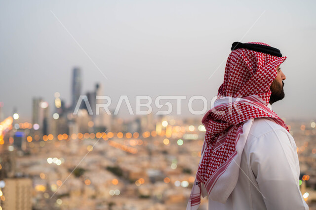 التنزه والتجول واستنشاق الهواء النقي في الخارج، الاعتزاز والفخر بالتقدم المعماري في السعودية، صورة من الخلف لرجل عربي خليجي سعودي يرتدي الشماغ و الزي السعودي التقليدي يقوم بتأمل معالم البلد، النظر الى التطور العمراني في المدينة، خلفية احياء مدينة الرياض.
