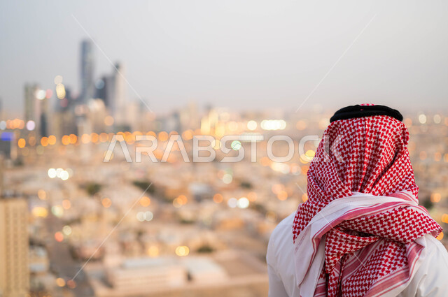 النظر الى التطور العمراني في المدينة، التنزه والتجول واستنشاق الهواء النقي في الخارج، الاعتزاز والفخر بالتقدم المعماري في السعودية، صورة من الخلف لرجل عربي خليجي سعودي يرتدي الشماغ و الزي السعودي التقليدي يقوم بتأمل معالم البلد، خلفية احياء مدينة الرياض.