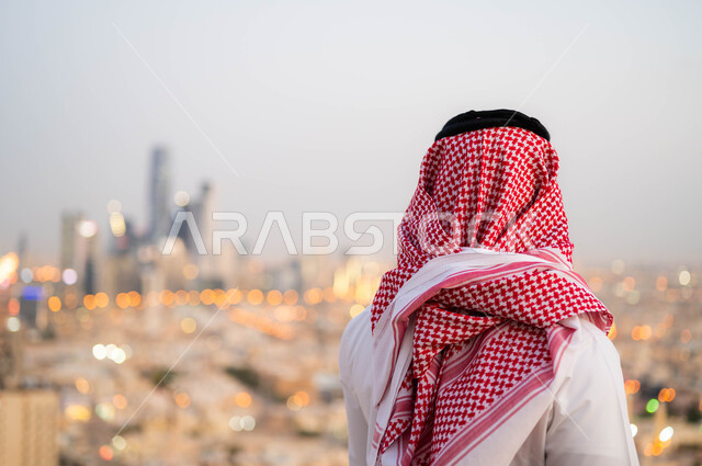 Walking around, hiking, breathing fresh air outside, taking pride in the architectural progress in Saudi Arabia, a picture from the back of a Saudi Arabian Gulf man wearing a shamagh and the traditional Saudi dress, contemplating the country's landmarks, 
