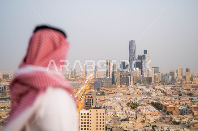 الاعتزاز والفخر بالتقدم المعماري، النمو و التطور العمراني في المدينة، التجول والتنزه واستنشاق الهواء النقي في الخارج، صورة من الخلف لرجل عربي خليجي سعودي يرتدي الشماغ و الزي التقليدي يقوم بالنظر إلى معالم البلد، خلفية الابراج المالية الاقتصادية في الرياض