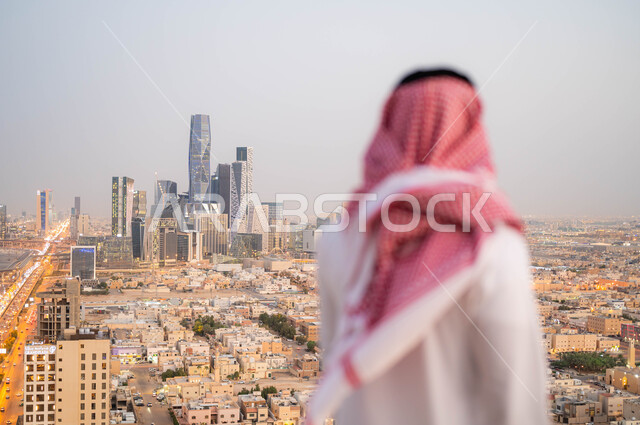 النمو و التطور العمراني في المدينة، الاعتزاز والفخر بالتقدم المعماري في السعودية، صورة من الخلف لرجل عربي خليجي سعودي يرتدي الشماغ و الزي التقليدي يقوم بالنظر إلى معالم البلد، خلفية ابراج الملك عبد الله المالية والاقتصادية في الرياض