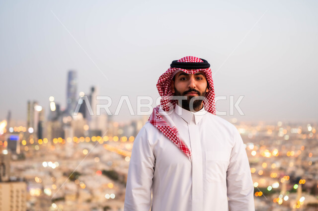 الاعتزاز والفخر بالتقدم المعماري في السعودية، التقدم والتطور العمراني في المدينة، التجول والتنزه واستنشاق الهواء النقي في الخارج، صورة مقربة لرجل عربي خليجي سعودي مبتسم يرتدي الشماغ و الزي السعودي التقليدي ينظر الى الكاميرا، خلفية احياء مدينة الرياض.