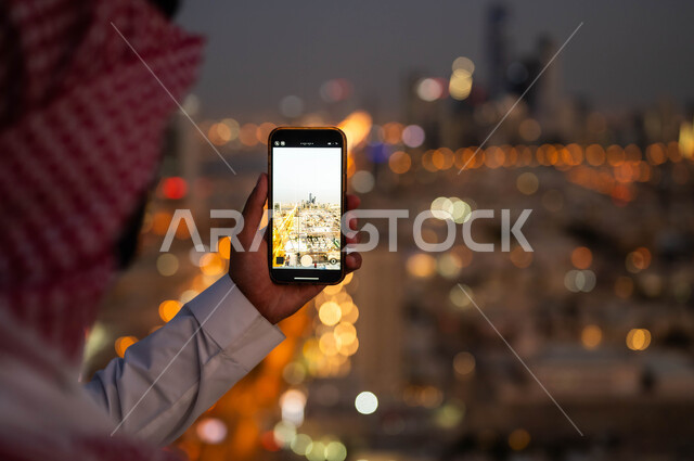 نشر معالم الرياض على وسائل التواصل الاجتماعي, مشاركة الثقافات الاخرى حضارة المملكة العربية السعودية، صورة مقربة من الخلف لشاب عربي خليجي سعودي يرتدي الشماغ والزي السعودي يحمل بيده الجوال، التقاط صور تذكارية للمدينة بإستخدام الهاتف المحمول ليلا