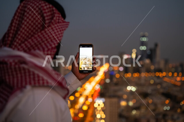 مشاركة الثقافات الاخرى حضارة المملكة العربية السعودية، نشر معالم الرياض على وسائل التواصل الاجتماعي, صورة مقربة من الخلف لشاب عربي خليجي سعودي يرتدي الشماغ والزي السعودي يحمل بيده الجوال، التقاط صور تذكارية للمدينة بإستخدام الهاتف المحمول ليلا