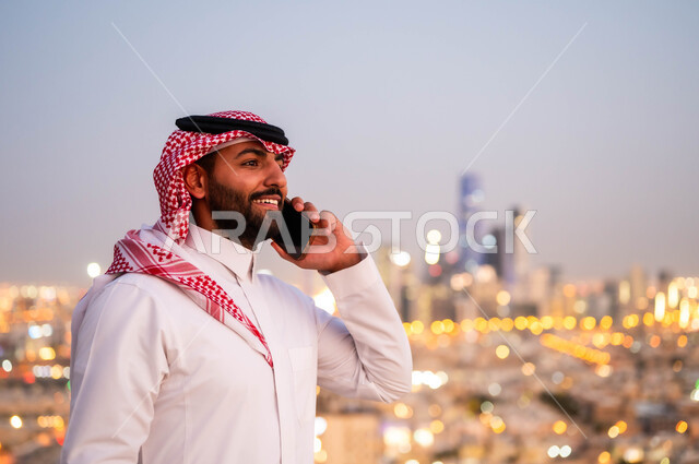  الدردشة و التواصل مع الاصدقاء، إجراء مكالمة هاتفية، صورة مقربة لشاب عربي خليجي سعودي مبتسم يرتدي الشماغ والزي السعودي التقليدي يقوم بالتحدث عبر الهاتف المحمول، الرد والاجابة على الاتصالات عبر الجوال، خلفية احياء ومباني مدينة الرياض.