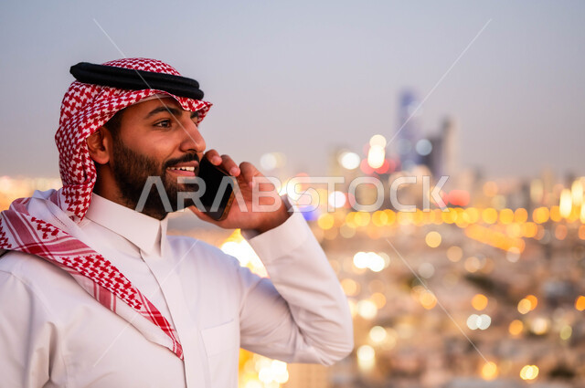  الرد والاجابة على الاتصالات عبر الجوال، صورة مقربة لشاب عربي خليجي سعودي مبتسم يرتدي الشماغ والزي السعودي التقليدي يقوم بالتحدث عبر الهاتف المحمول، الدردشة و التواصل مع الاصدقاء، إجراء مكالمة هاتفية، خلفية احياء ومباني مدينة الرياض.