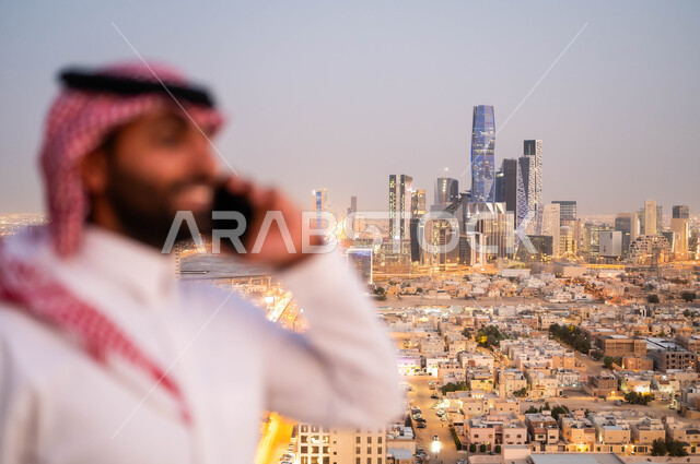 إجراء مكالمة هاتفية عبر الجوال، صورة مقربة لشاب عربي خليجي سعودي مبتسم يرتدي الشماغ والزي التقليدي يقوم بالتحدث عبر الهاتف المحمول، الدردشة و التواصل مع الاصدقاء، الرد والاجابة على الاتصالات، خلفية ابراج الملك عبد الله الاقتصادية والمالية في الرياض