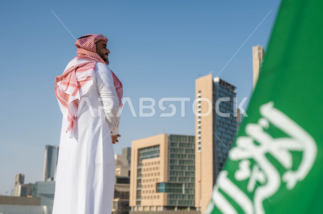 الفخر والاعتزاز بامتلاك الهوية الوطنية، صورة مقربة لعلم المملكة العربية السعودية في ذكرى اليوم الوطني السعودي 23 سبتمبر، رجل عربي خليجي سعودي يرتدي الشماغ والثوب التقليدي يقوم بمشاهدة أبراج وناطحات سحاب مدينة الرياض في وضح النهار، الاحتفال بيوم العلم 11 مارس