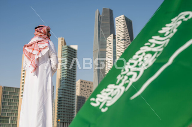 الفخر والاعتزاز بامتلاك الهوية الوطنية، صورة مقربة لعلم المملكة العربية السعودية في ذكرى اليوم الوطني السعودي 23 سبتمبر، رجل عربي خليجي سعودي يرتدي الشماغ والثوب التقليدي يقوم بمشاهدة أبراج وناطحات سحاب مدينة الرياض في وضح النهار، الاحتفال بيوم العلم 11 مارس