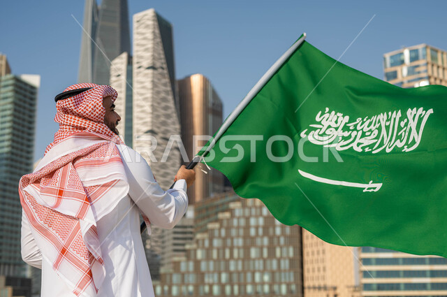 اليوم الوطني 8 ربيع الأول, الفخر و الاعتزاز بالانتماء للمملكة, ذكرى العيد الوطني السعودي, صورة من الخلف لرجل عربي سعودي خليجي يرتدي الشماغ والثوب التقليدي يحمل بيده علم السعودية للاحتفال بيوم العلم 11مارس, خلفية أبراج مدينة الرياض في وضح النهار