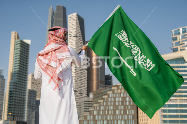 استذكار التاريخ والهوية الوطنية, الاحتفال بيوم العلم 11 مارس, ذكرى اليوم الوطني السعودي 23 سبتمبر, صورة من الخلف لرجل عربي خليجي سعودي يرتدي الثوب السعودي يحمل بيده العلم الوطني, العيد الوطني السعودي, خلفية ابراج الملك عبد الله