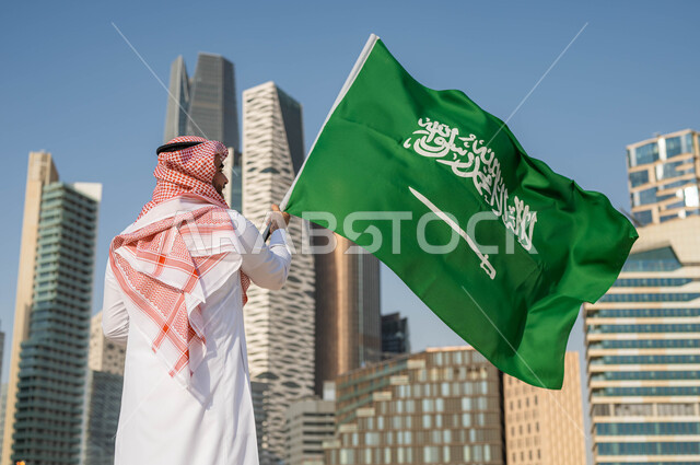 استذكار التاريخ والهوية الوطنية, الاحتفال بيوم العلم 11 مارس, ذكرى اليوم الوطني السعودي 23 سبتمبر, صورة من الخلف لرجل عربي خليجي سعودي يرتدي الثوب السعودي يحمل بيده العلم الوطني, العيد الوطني السعودي, خلفية ابراج الملك عبد الله