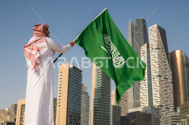 الاحتفال بيوم العلم 11 مارس, استذكار التاريخ والهوية الوطنية, ذكرى اليوم الوطني السعودي 23 سبتمبر, صورة من الخلف لرجل عربي خليجي سعودي يرتدي الثوب السعودي يحمل بيده العلم الوطني, العيد الوطني السعودي, خلفية ابراج الملك عبد الله