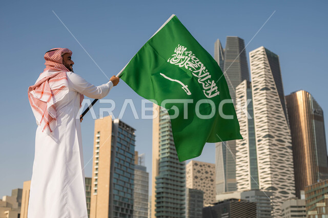 الاحتفال بيوم العلم 11 مارس, استذكار التاريخ والهوية الوطنية, ذكرى اليوم الوطني السعودي 23 سبتمبر, صورة من الخلف لرجل عربي خليجي سعودي يرتدي الثوب السعودي يحمل بيده العلم الوطني, العيد الوطني السعودي, خلفية ابراج الملك عبد الله