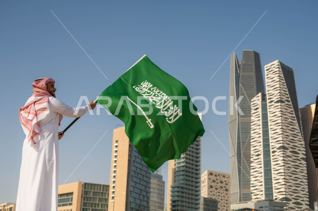 الاحتفال بيوم العلم 11 مارس, استذكار التاريخ والهوية الوطنية, ذكرى اليوم الوطني السعودي 23 سبتمبر, صورة من الخلف لرجل عربي خليجي سعودي يرتدي الثوب السعودي يحمل بيده العلم الوطني, العيد الوطني السعودي, خلفية ابراج الملك عبد الله