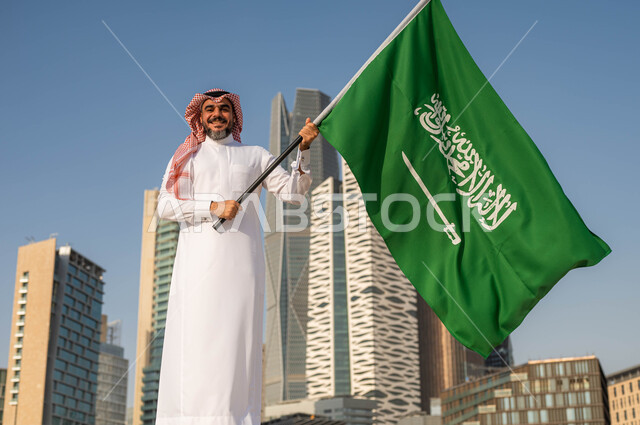 يوم العلم 11مارس، الاحتفال بذكرى اليوم الوطني السعودي 8 ربيع الأول, الفخر و الاعتزاز بالانتماء للمملكة, رجل عربي سعودي خليجي مبتسم يرتدي الشماغ والثوب التقليدي يحمل بيده علم السعودية ينظر الى الكاميرا, خلفية أبراج مدينة الرياض في وضح النهار