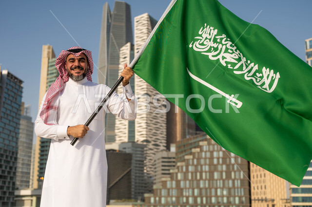 Flag Day, March 11, celebration of the anniversary of the Saudi National Day, Rabi’ al-Awwal 8, pride and pride in belonging to the Kingdom, a close-up photo of a smiling Saudi Gulf Arab man wearing a shemagh and traditional thobe, holding the Saudi flag in his hand, looking at the camera, background of the towers of the city of Riyadh in broad daylight