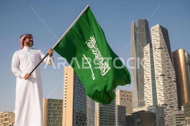 خلفية مركز الملك عبدلله المالي وضح النهار، الفخر والاعتزاز بالانتماء للمملكة, يوم العلم 11مارس، الاحتفال بذكرى اليوم الوطني السعودي 8 ربيع الأول, رجل عربي سعودي خليجي مبتسم يرتدي الشماغ والثوب التقليدي يحمل بيده علم السعودية ينظر اليه بحب واحترام