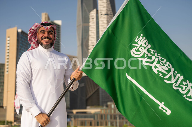الفخر والاعتزاز بالانتماء للمملكة, يوم العلم 11مارس، الاحتفال بذكرى اليوم الوطني السعودي 8 ربيع الأول, صورة مقربة لرجل عربي سعودي خليجي مبتسم يرتدي الشماغ والثوب التقليدي يحمل بيده علم السعودية ينظر الى الكاميرا, خلفية أبراج مدينة الرياض وضح النهار