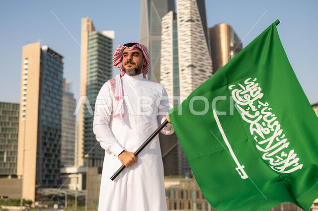 ايماءات  الفخر والاعتزاز بالانتماء للمملكة, يوم العلم 11مارس، الاحتفال باليوم الوطني 8 ربيع الأول, ذكرى العيد الوطني السعودي, رجل عربي سعودي خليجي مبتسم يرتدي الشماغ والثوب التقليدي يحمل بيده علم السعودية, خلفية مركز الملك عبدلله المالي وضح النهار