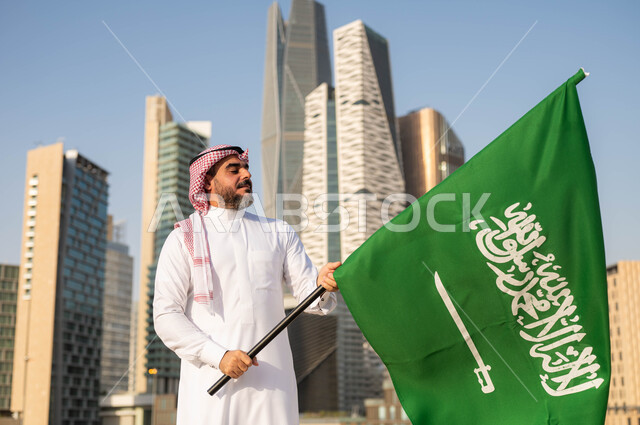ايماءات  الفخر والاعتزاز بالانتماء للمملكة, يوم العلم 11مارس، الاحتفال باليوم الوطني 8 ربيع الأول, ذكرى العيد الوطني السعودي, رجل عربي سعودي خليجي مبتسم يرتدي الشماغ والثوب التقليدي يحمل بيده علم السعودية, خلفية مركز الملك عبدلله المالي وضح النهار
