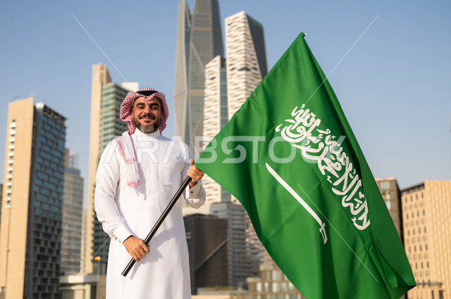 ايماءات  الفخر والاعتزاز بالانتماء للمملكة, يوم العلم 11مارس، الاحتفال باليوم الوطني 8 ربيع الأول, ذكرى العيد الوطني السعودي, رجل عربي سعودي خليجي مبتسم يرتدي الشماغ والثوب التقليدي يحمل بيده علم السعودية, خلفية مركز الملك عبدلله المالي وضح النهار