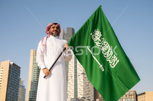 يوم العلم 11مارس، الاحتفال باليوم الوطني 8 ربيع الأول, الفخر و الاعتزاز بالانتماء للمملكة, ذكرى العيد الوطني السعودي, رجل عربي سعودي خليجي مبتسم يرتدي الشماغ والثوب التقليدي يحمل بيده علم السعودية ينظر الى الكاميرا, خلفية أبراج مدينة الرياض في وضح النهار