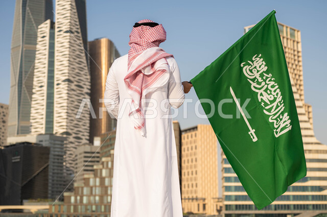 اليوم الوطني 8 ربيع الأول, الفخر و الاعتزاز بالانتماء للمملكة, ذكرى العيد الوطني السعودي, صورة من الخلف لرجل عربي سعودي خليجي يرتدي الشماغ والثوب التقليدي يحمل بيده علم السعودية للاحتفال بيوم العلم 11مارس, خلفية أبراج مدينة الرياض في وضح النهار