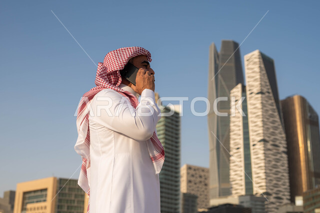 الاجابة والرد على الاتصالات باستخدام الجوال، صورة مقربة لرجل عربي خليجي سعودي مبتسم يرتدي الشماغ والثوب التقليدي يقوم بالتحدث عبر الهاتف المحمول في وضح النهار، الدردشة و التواصل مع الاصدقاء، إجراء مكالمة هاتفية، خلفية مركز الملك عبدلله المالي