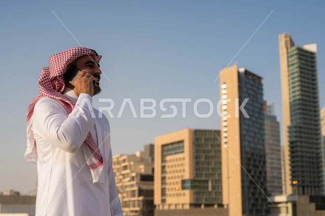 الدردشة و التواصل مع الاصدقاء، صورة مقربة لرجل عربي خليجي سعودي مبتسم يرتدي الشماغ والثوب التقليدي يقوم بالتحدث عبر الهاتف المحمول في وضح النهار، الاجابة والرد على الاتصالات باستخدام الجوال، إجراء مكالمة هاتفية، خلفية أبراج مدينة الرياض