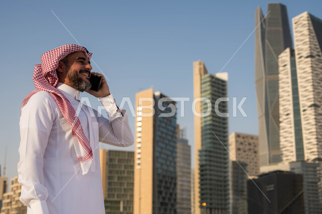 الرد والاجابة على الاتصالات باستخدام الجوال، صورة مقربة لرجل عربي خليجي سعودي مبتسم يرتدي الشماغ والثوب التقليدي يقوم بالتحدث عبر الهاتف المحمول في وضح النهار، الدردشة و التواصل مع الاصدقاء، إجراء مكالمة هاتفية، خلفية مركز الملك عبدلله المالي