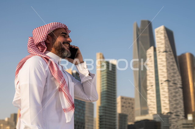 الرد والاجابة على الاتصالات باستخدام الجوال، صورة مقربة لرجل عربي خليجي سعودي مبتسم يرتدي الشماغ والثوب التقليدي يقوم بالتحدث عبر الهاتف المحمول في وضح النهار، الدردشة و التواصل مع الاصدقاء، إجراء مكالمة هاتفية، خلفية مركز الملك عبدلله المالي