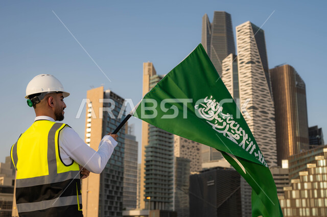 الاحتفال بيوم العلم 11 مارس, دور المهندسيين في التطور العمراني, إحياء ذكرى اليوم الوطني السعودي في 23 سبتمبر, صورة من الخلف لمهندس عربي سعودي خليجي يرتدي سترة الحماية والخوذة البيضاء يمسك علم المملكة بفخر واعتزاز, خلفية مركز الملك عبدلله المالي وضح النهار