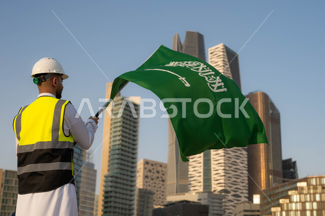 Commemoration of the Saudi National Day on September 23, the role of engineers in urban development, celebration of Science Day on March 11, a picture from behind of a smiling Saudi Arabian Gulf engineer wearing a protection vest and white helmet holding the Kingdom’s flag with pride, background of the towers of the city of Riyadh in broad daylight