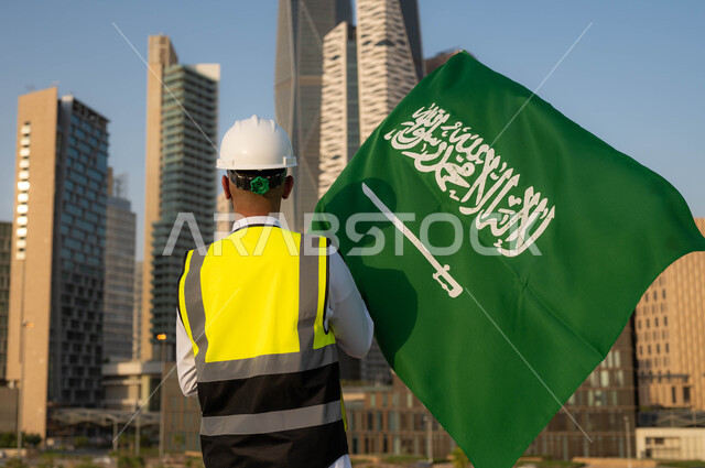 إحياء ذكرى اليوم الوطني السعودي في 23 سبتمبر, دور المهندسيين في التطور العمراني, الاحتفال بيوم العلم 11 مارس, صورة من الخلف لمهندس عربي سعودي خليجي يرتدي سترة الحماية والخوذة البيضاء يمسك علم المملكة بفخر واعتزاز, خلفية أبراج مدينة الرياض وضح النهار