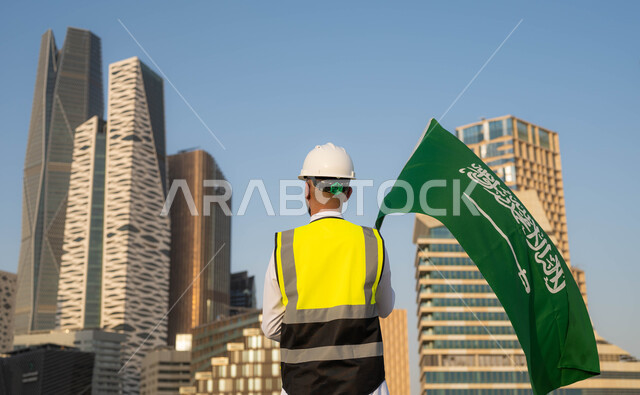 دور المهندسيين في التطور العمراني, الاحتفال بيوم العلم 11 مارس, إحياء ذكرى اليوم الوطني السعودي في 23 سبتمبر, صورة من الخلف لمهندس عربي سعودي خليجي يرتدي سترة الحماية والخوذة البيضاء يمسك علم المملكة بفخر واعتزاز, خلفية مركز الملك عبدلله المالي وضح النهار