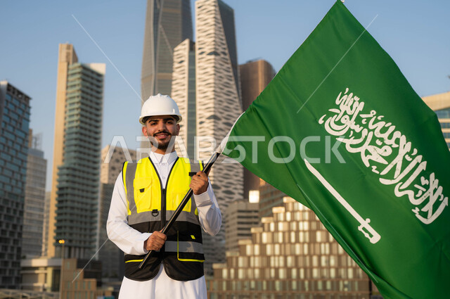 دور المهندسيين في التطور العمراني, الاحتفال بيوم العلم 11 مارس, إحياء ذكرى اليوم الوطني السعودي في 23 سبتمبر, مهندس معماري عربي سعودي خليجي مبتسم يرتدي سترة الحماية والخوذة البيضاء يمسك علم المملكة بفخر واعتزاز, خلفية مركز الملك عبدلله المالي وضح النهار