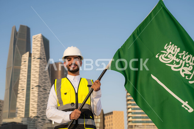 دور المهندسيين في التطور العمراني, الاحتفال بيوم العلم 11 مارس, إحياء ذكرى اليوم الوطني السعودي في 23 سبتمبر, صورة مقربة لمهندس عربي سعودي خليجي مبتسم يرتدي سترة الحماية والخوذة البيضاء يمسك علم المملكة بفخر واعتزاز, خلفية مركز الملك عبدلله المالي وضح النهار