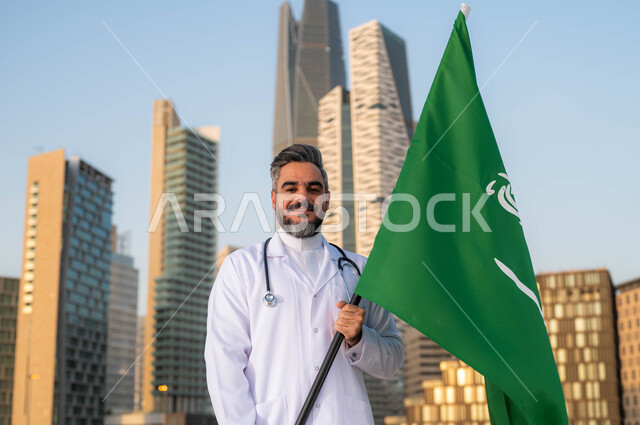 تقديم الرعاية الصحة لابناء الوطن، الاحتفالات بذكرى اليوم الوطني السعودي 23 سبتمبر، صورة مقربة لطبيب عربي خليجي سعودي يرتدي المعطف الأبيض يمسك بيده علم السعودية بإيماءات الفخر والاعتزاز, يوم العلم 11 مارس، خلفية مركز الملك عبدلله المالي وضح النهار